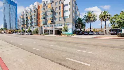 A modern apartment complex exterior with palm trees, balconies, and street parking under a bright blue sky.