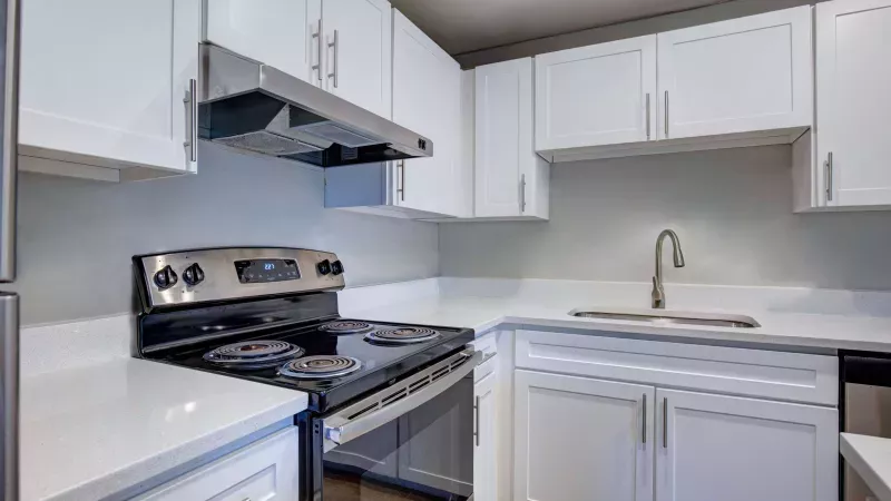 Contemporary kitchen with sleek white cabinetry, stainless steel appliances, and an electric stovetop. The space features modern brushed nickel hardware, quartz-style countertops, and a gooseneck faucet above a deep single-basin sink. The neutral gray backsplash and clean design offer a bright and minimalist aesthetic.