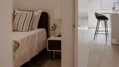 A bedroom view through an open doorway, showing a neatly styled bed and a glimpse of the modern kitchen in the background.