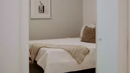 A cozy bedroom with a minimalist design, featuring a neatly made bed with soft beige bedding, decorative pillows, and a framed art piece on the wall.