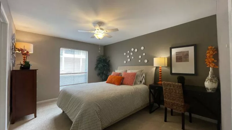 A beautifully designed bedroom with a large bed, decorative pillows, a ceiling fan, and a well-lit workspace, creating a comfortable and inviting atmosphere.