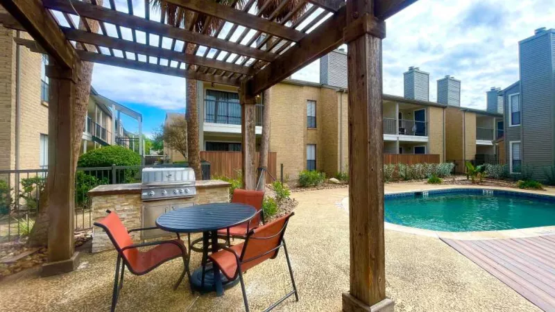 A shaded pergola with a built-in grilling station and seating area, overlooking the pool at The Oaks of Westchase Apartments.