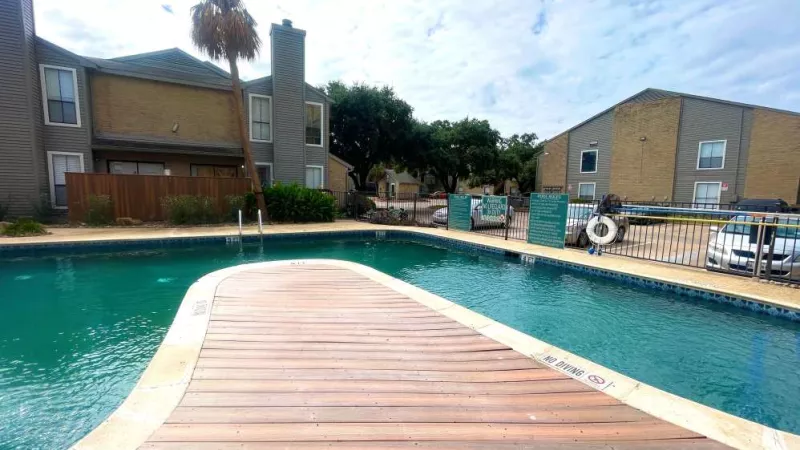 A scenic view of the swimming pool at The Oaks of Westchase Apartments, featuring a stylish wooden bridge and lush greenery, surrounded by modern apartment buildings.