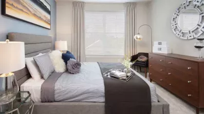 A sophisticated primary bedroom featuring a gray upholstered bed, elegant furnishings, and a large window for natural brightness.