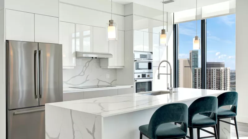 A modern white kitchen with marble countertops, stainless steel appliances, and large windows overlooking a city skyline.