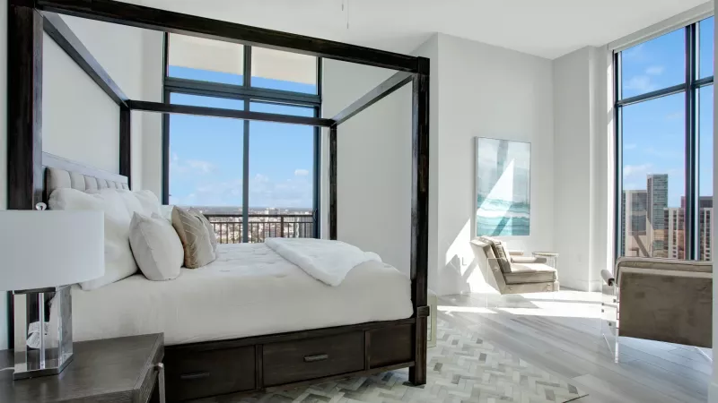 A spacious bedroom featuring a dark wood canopy bed, plush white bedding, and floor-to-ceiling windows with city views.