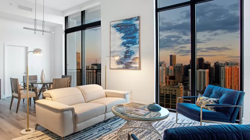 A stylish living room with a cream-colored couch, blue accent chairs, and floor-to-ceiling windows revealing a stunning sunset over the city skyline.