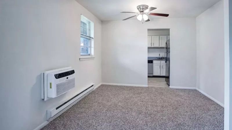 A spacious living area with neutral walls, plush carpeting, a ceiling fan, and a window unit air conditioner, leading to a modern kitchen with white cabinetry.