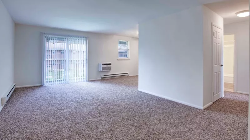A well-lit apartment interior featuring an expansive living space, plush carpeting, and access to a private balcony through a sliding glass door.