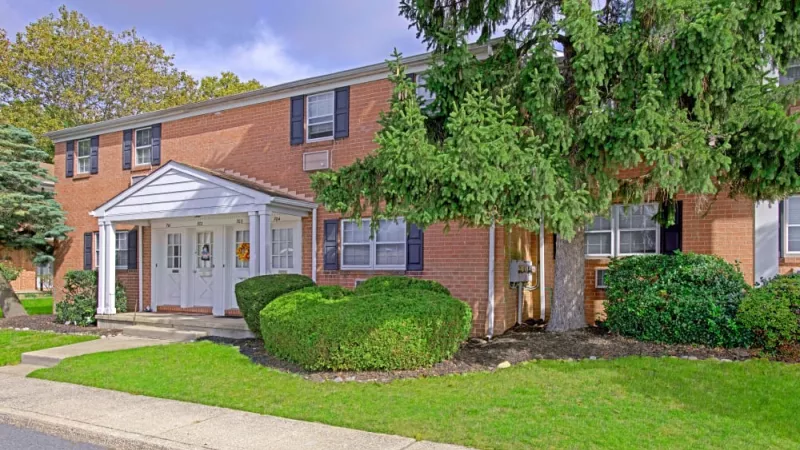 A welcoming entrance to Silver Lake Manor Apartments, featuring well-manicured shrubs, a white-trimmed door, and a charming porch under a large pine tree.