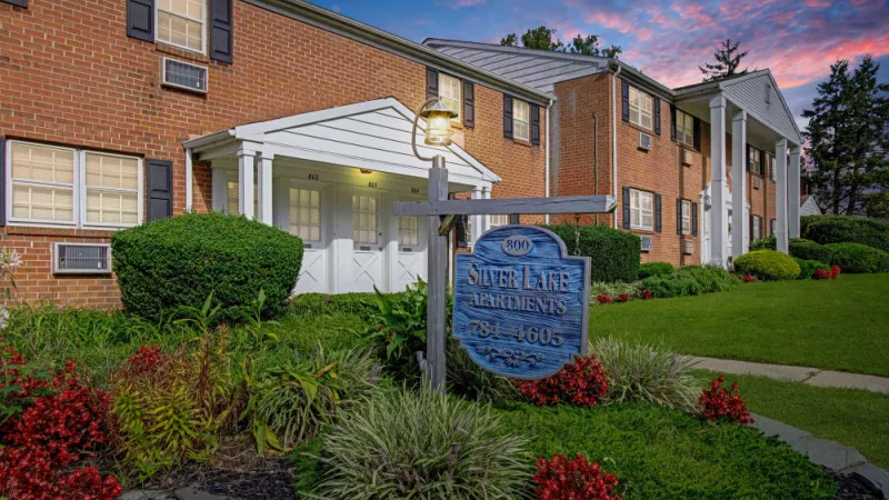 Silver Lake Manor Apartments entrance at dusk, featuring a warmly lit sign, lush landscaping, and a brick colonial-style facade.