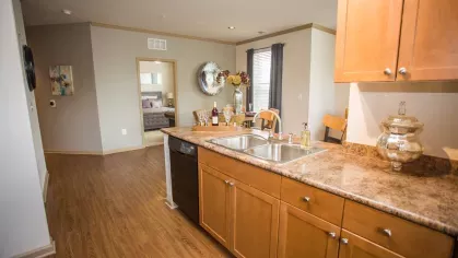 A well-lit kitchen with wood cabinetry, granite-style countertops, and a dual sink overlooking the dining and living area. The open layout maximizes space and functionality.