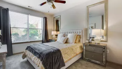 A bright and airy bedroom featuring a plush, tufted bed with soft beige and gray bedding. Large mirrors, stylish nightstands, and warm lighting create a luxurious and inviting atmosphere.
