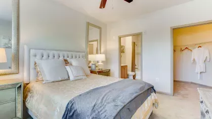 A well-appointed bedroom with a tufted headboard, neutral bedding, and mirrored nightstands, leading to an open walk-in closet and en-suite bathroom.