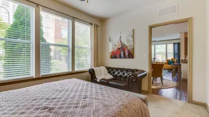A spacious bedroom with a modern gray bed, large windows, and an adjacent sitting area featuring a leather couch, leading into the open living and dining space.