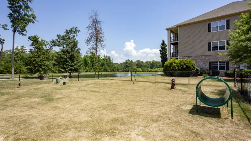 Fenced dog park at Lauren Ridge featuring agility equipment and open space, set against a backdrop of trees and a scenic pond.