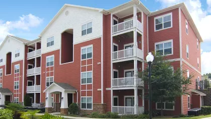 Red and white three-story apartment building with balconies at Reed Creek Apartments surrounded by landscaped greenery.
