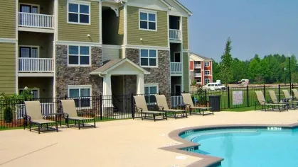 Three-story green and beige apartment building beside pool with tan lounge chairs