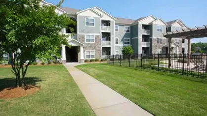 Green apartment building exterior with manicured lawn, fenced courtyard, and paved walkway.