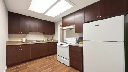 Modern galley-style kitchen at Astorwood Apartments featuring dark wood cabinetry, white appliances, and wood-look flooring under bright overhead lighting.