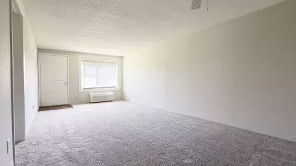 Large unfurnished living room with plush carpet, white ceiling fan, and wide front window providing natural light at Astorwood Apartments.