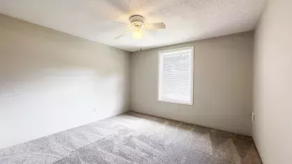 Empty primary bedroom with light gray carpet, white ceiling fan, and single large window allowing for natural light at Astorwood Apartments.