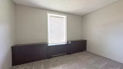 Unfurnished second bedroom with built-in dark wood storage, soft carpet, and a centered window with blinds at Astorwood Apartments.