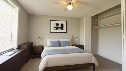 Virtually staged bedroom with a soft neutral bed, gold reading lamps, built-in closet with open shelving, and large window with natural light at Astorwood Apartments.