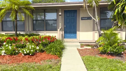 Exterior view of Astorwood Apartments leasing office with a blue door, beige siding, colorful flower beds, and palm trees.