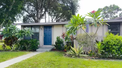 Front view of an Astorwood apartment home with blue door, large windows, and vibrant tropical landscaping including flowering bushes and palm trees.