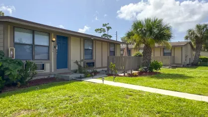 Exterior of a ground-level apartment at Astorwood Apartments featuring private fenced patio, blue door, and palm trees in a green courtyard setting.