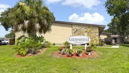 Astorwood Apartments community sign surrounded by flower beds, palm trees, and lush green landscaping in a sunny Florida setting.