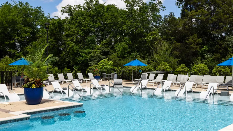 Poolside lounge chairs and blue umbrellas at Tapestry Turkey Creek with a view of lush greenery.
