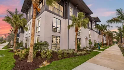 Exterior view of Tropia Wellen Park apartment building with modern design, palm trees, and well-manicured landscaping at sunset.