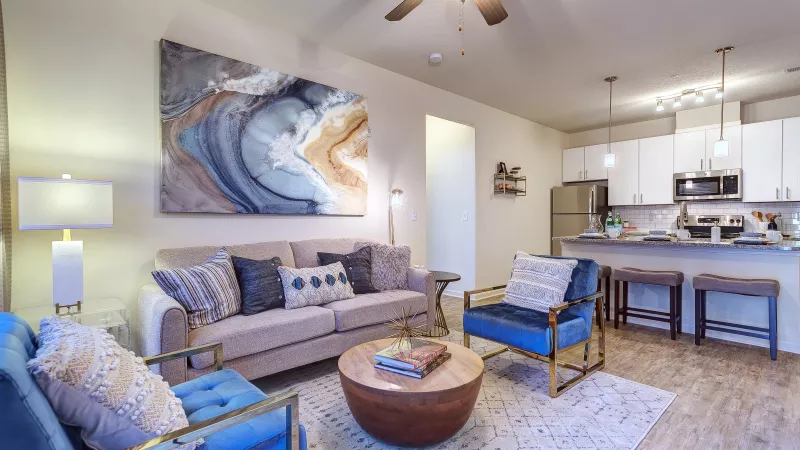 Stylish apartment living room with a beige sofa, blue accent chairs, modern artwork, and an open kitchen with bar seating and stainless-steel appliances.