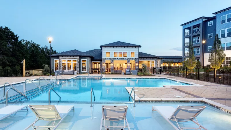 Expansive outdoor swimming pool with built-in lounge chairs on a sun shelf, surrounded by a spacious deck. In the background, a modern clubhouse with large windows glows warmly at dusk, framed by landscaped grounds and contemporary apartment buildings.