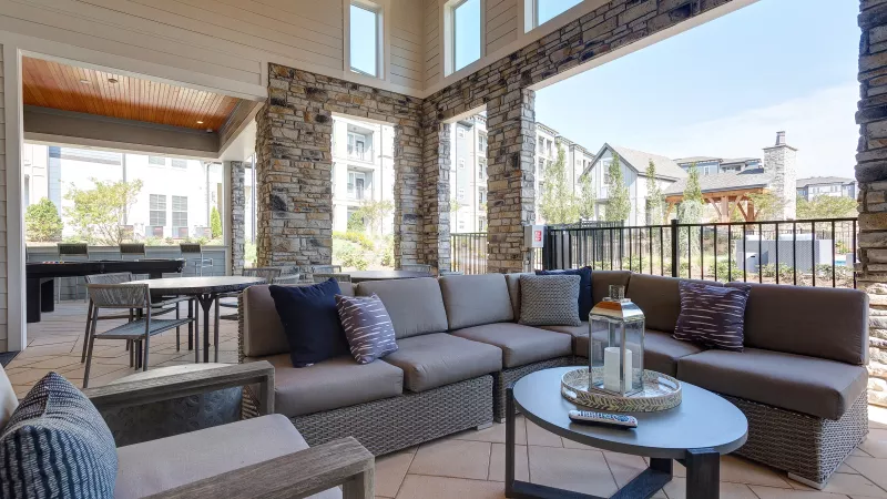 Covered outdoor seating area with stone columns, cushioned wicker furniture, accent pillows, and a modern coffee table with lantern décor, overlooking the apartment community.