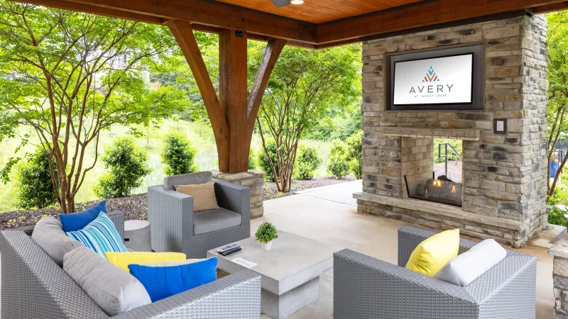 Covered outdoor lounge featuring modern wicker seating, colorful accent pillows, a stone fireplace with mounted TV, and views of landscaped greenery.