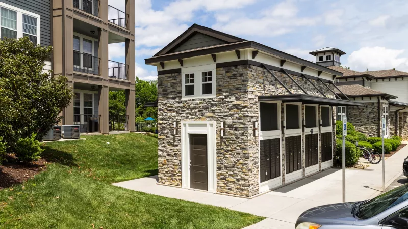 Community mail center with stone exterior and covered mailboxes, located next to modern apartment buildings and landscaped green space.