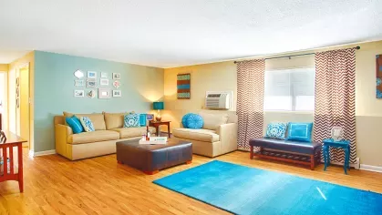 Bright and colorful living room with wood-style flooring, beige sofas, and bold turquoise accents. The space features patterned curtains, a blue accent wall with framed art, a large blue area rug, and decorative pillows. Natural light enters through the window, creating a cheerful and inviting atmosphere.