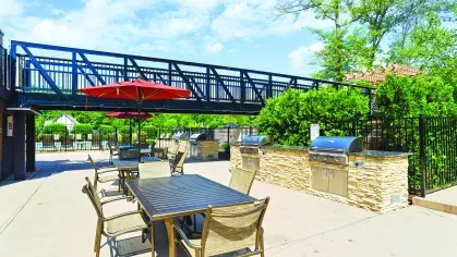 Outdoor grilling and dining area with stainless steel barbecue stations, stone countertops, tables with umbrellas, and a pedestrian bridge overhead, surrounded by greenery.