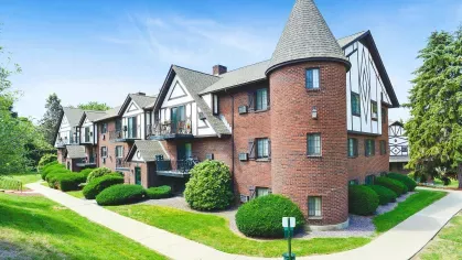 Charming brick apartment building with Tudor-style accents, gabled roofs, and a rounded turret feature. The property has well-maintained landscaping with green lawns, neatly trimmed shrubs, and mature trees under a bright blue sky.