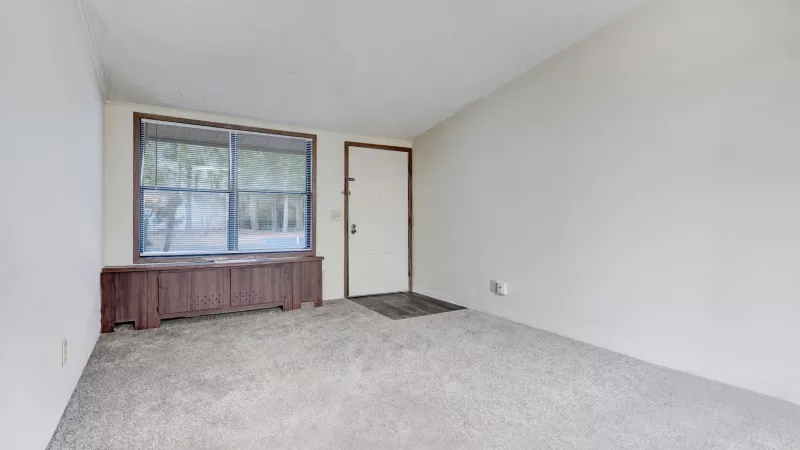 Bright living space with large front window, neutral carpeting, and front entry door.