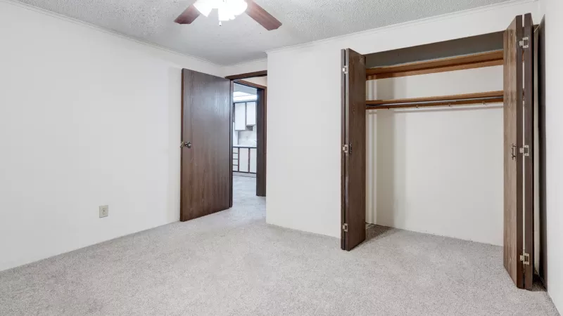 Carpeted bedroom with ceiling fan, double-door closet, and view into kitchen area.