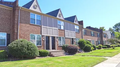 Exterior view of Maple Bay Apartment Townhomes featuring red brick facades, manicured shrubs, and neatly trimmed lawns under a bright blue sky.
