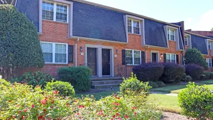 Maple Bay Apartment Townhomes with red brick walls, dark mansard-style roofs, and colorful shrubs in full bloom.