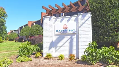 Secondary entrance monument for Maple Bay Apartment Townhomes with white brick sign, blue lettering, wood pergola detail, and vibrant surrounding greenery.