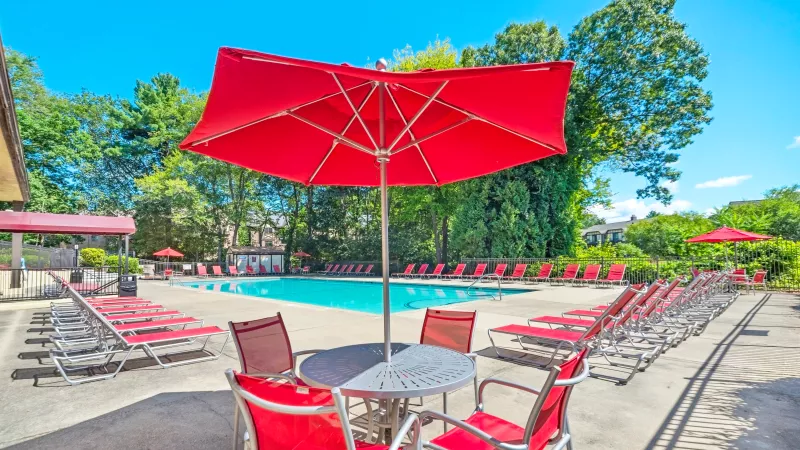 Outdoor swimming pool area with red lounge chairs and red umbrellas, featuring a shaded table and chairs in the foreground, surrounded by trees on a sunny day.