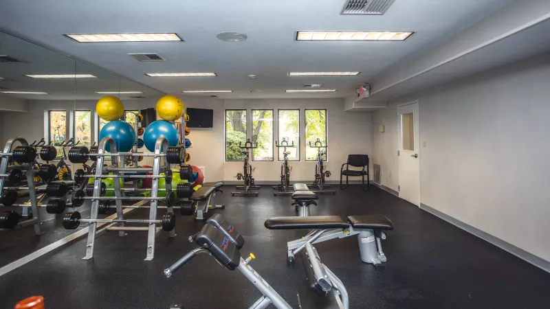 Small fitness center with free weights, weight benches, exercise balls, and stationary bikes. The room has large mirrors, bright overhead lighting, and windows providing natural light.