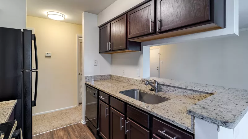 Modern kitchen with dark wood cabinets, granite countertops, stainless steel sink, and black appliances. The kitchen features wood-style flooring, neutral walls, and recessed lighting, with an open pass-through to the adjacent living area.